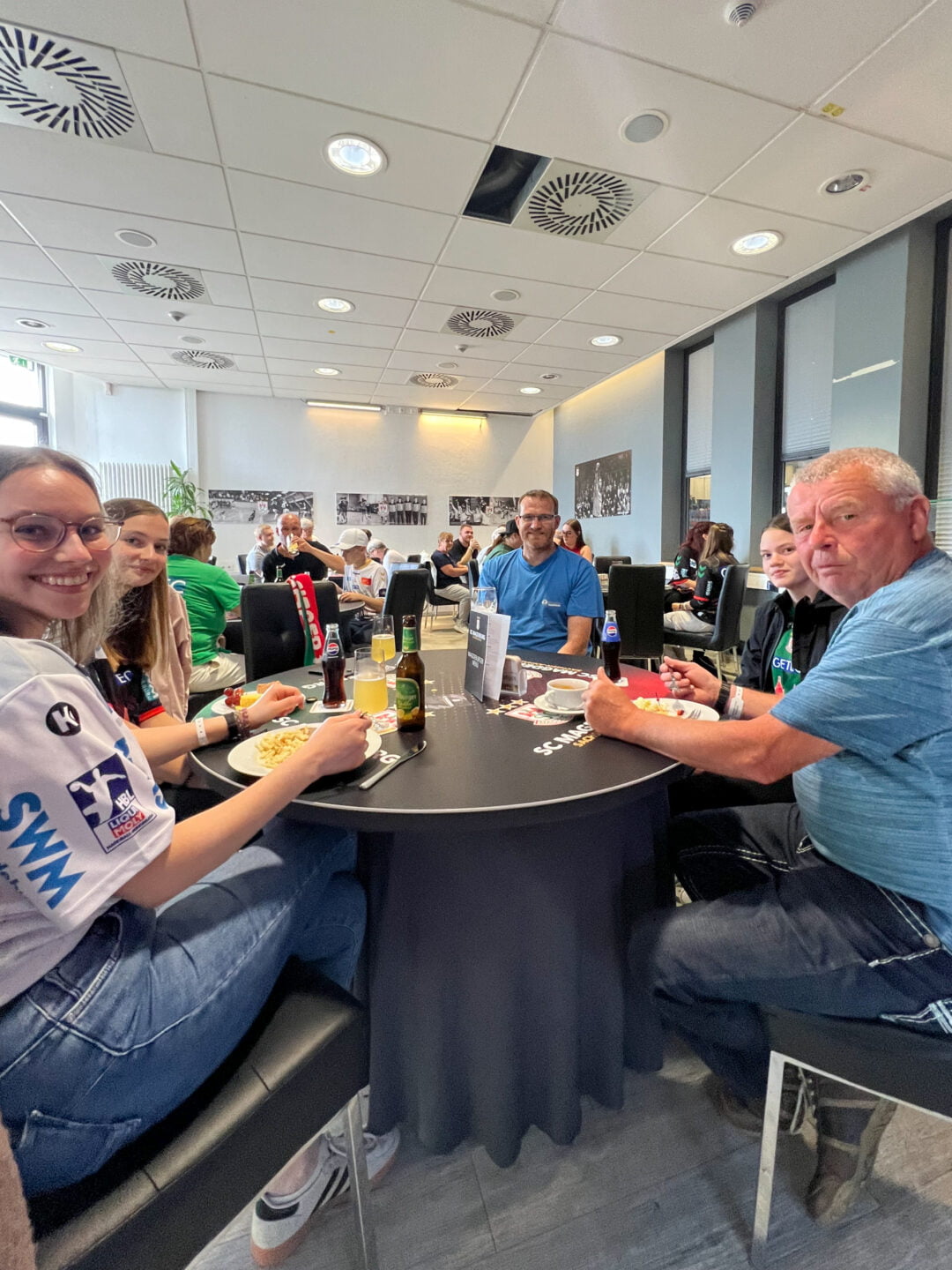 5 Personen sitzen an einem Tisch in der Klaus-Miesner-Lounge der GETEC Arena und schauen in die Kamera. Sie haben Getränke oder Essen vor sich stehen. Im Hintergrund sitzen weitere Menschen an Tischen.