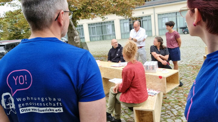 Vorstand Oliver Hornemann steht links im Bildvordergrund und schaut auf Teilnehmer einer Pressekonferenz, die auf großen Holzzylindern sitzen.