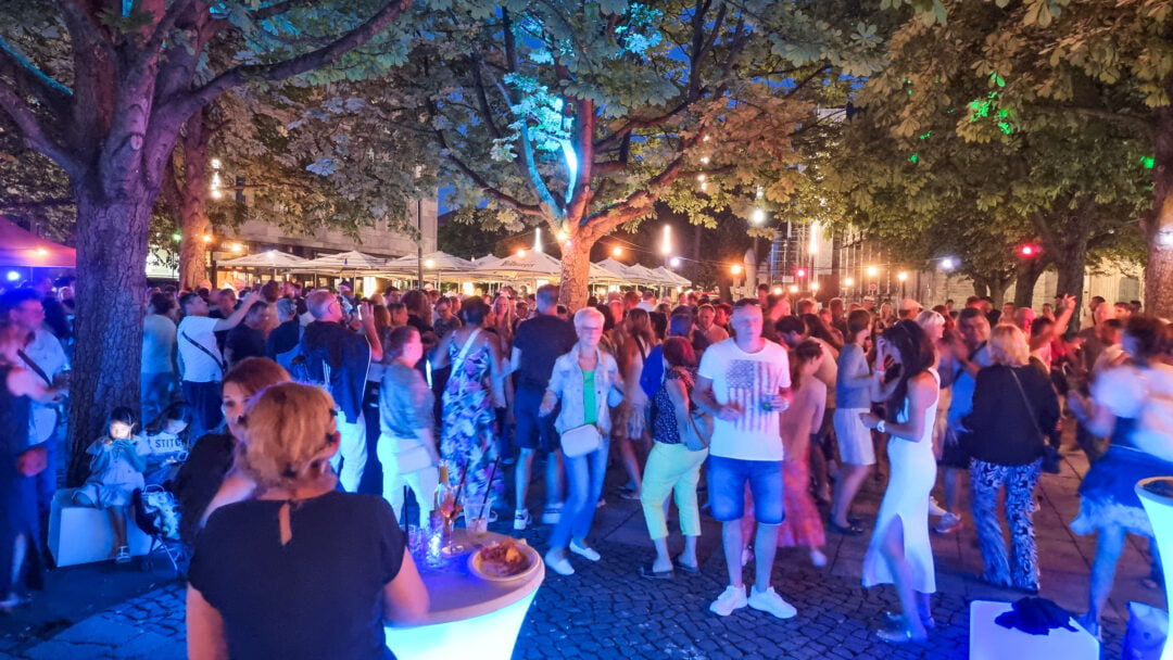 Eine große Gruppe von Menschen tanzen unter Kastanienbäumen im Abendlicht. Im Hintergrund der Magdeburger Dom.