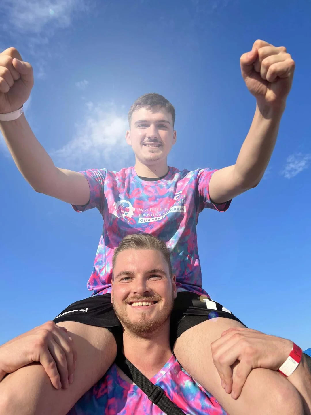 Ein jubelnder Mann auf den Schultern eines anderen unter blauem Himmel in Sportkleidung.