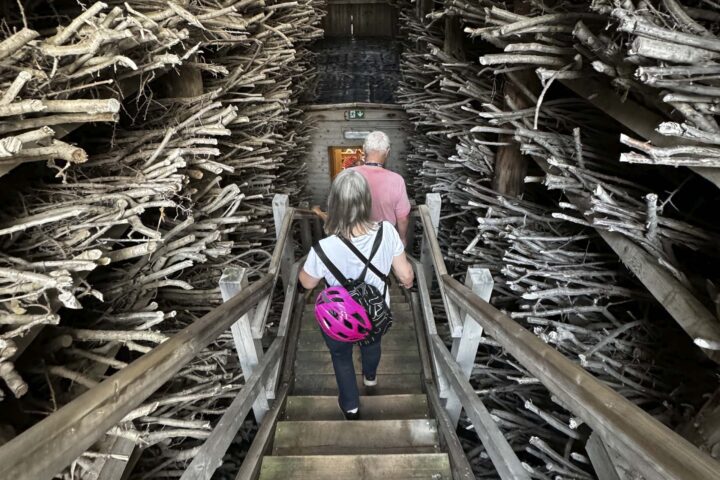 Zwei Menschen steigen eine Holztreppe hinunter. Eine Frau davon trägt einen pinken Fahrradhelm auf dem Rücken.