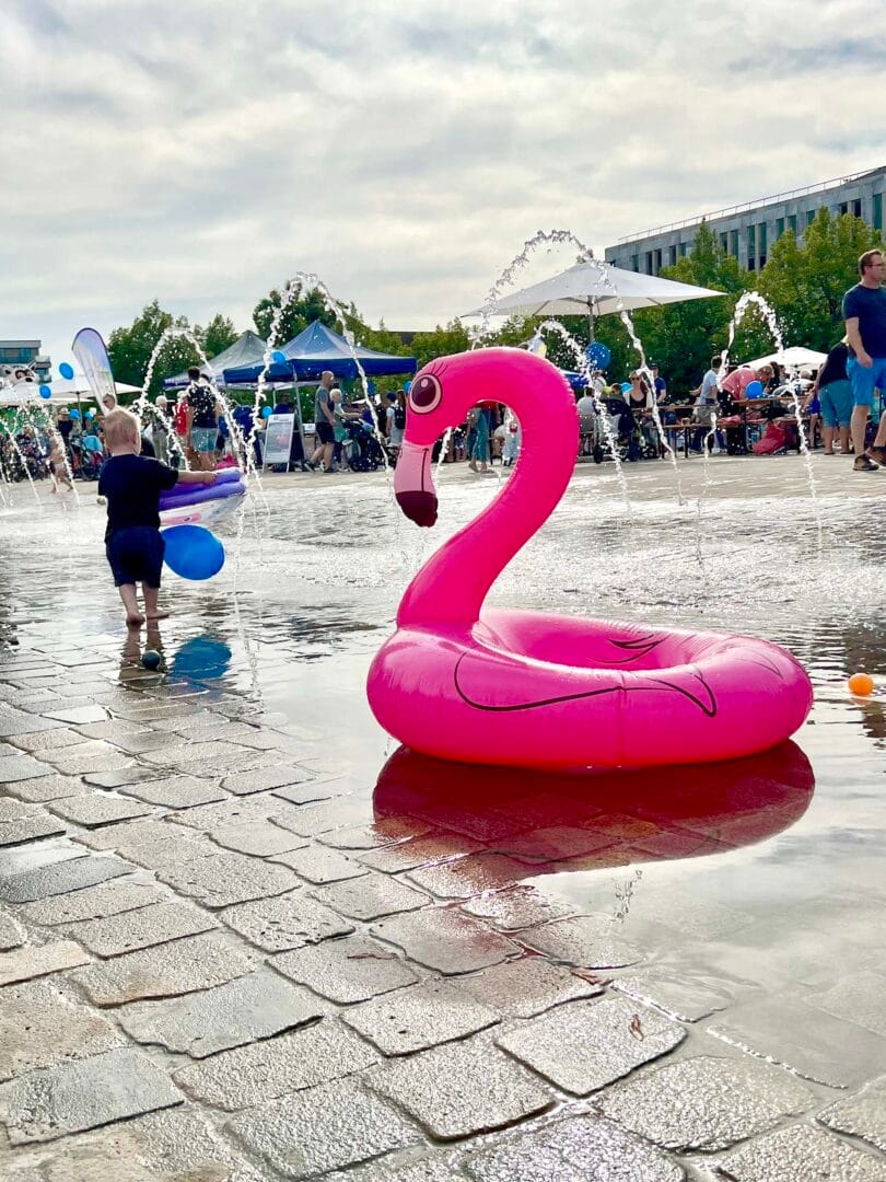 Ein pinker aufblasbarer Flamingo-Schwimmring liegt im Wasser auf einem Platz. Im Hintergrund eine Menschenmenge