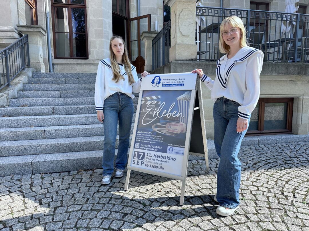 zwei junge Frauen in weißen Blusen stehen neben einem Aufsteller und lächeln in die Kamera.