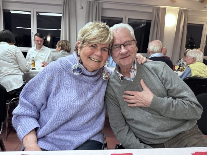 Ein Mann und eine Frau lächeln in die Kamera, weitere Menschen sitzen im Hintergrund an einem Tisch