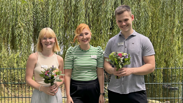 Zwei junge Frauen und ein junger Mann stehen vor einer Trauerweide auf einer Wiese.