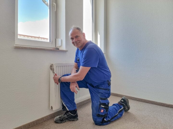 Ein Mann mit blauer Arbeitskleidung kniet vor einer Heizung, repariert sie und lächelt in die Kamera.