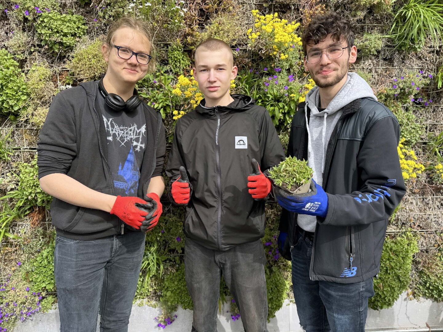Drei junge Männer in schwarzen Jacken schauen in die kamera und lächeln. Einer davon hat eine Pflanzen in der Hand, der andere junge Mann zeigt beide daumen nach oben.