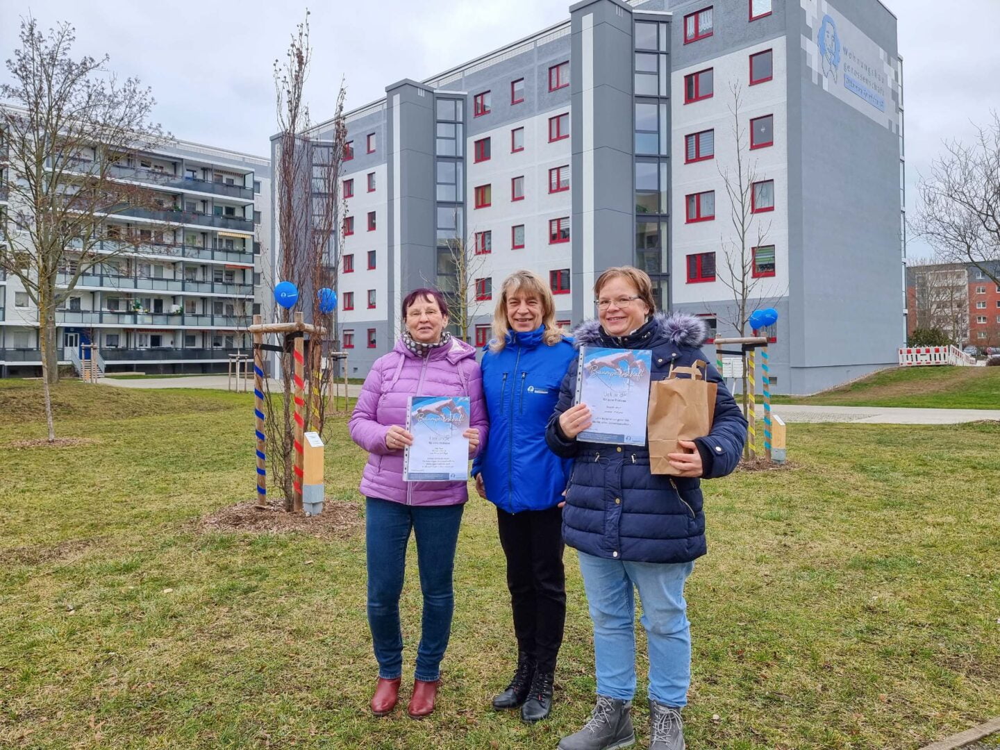Drei Frauen lächeln in die Kamera und halten eine Urkunde in der Hand im Hintergrund ein Baum und ein Wohnhaus mit Balkonen ©Anja Jänichen