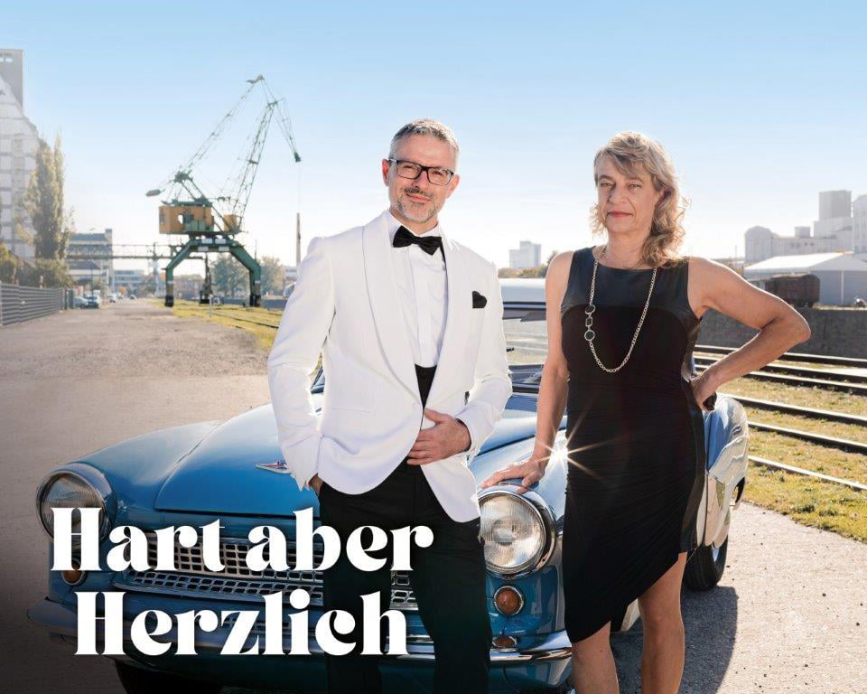 Eine Frau im schwarzen Abendkleid und ein Mann mit weißem Sakko, stehen vor einem alten blauen Auto. Davor steht in weißer Schrift, Hart aber Herzlich.