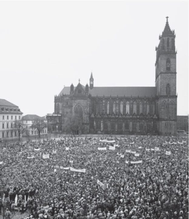 Das Bild zeigt eine große Menschenmenge, die sich vor dem Magdeburger Dom versammelt hat. Viele Personen halten Transparente und Schilder hoch, was auf eine Demonstration oder Kundgebung hinweist. Der Dom bildet den Hintergrund des Geschehens.