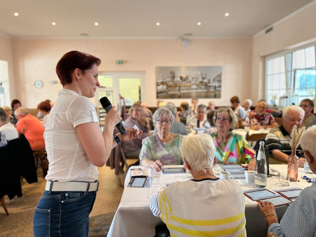 Frau in einer weißer Bluse steht vor einem Tisch mit vielen Senioren und hält ein Mirkrofon in der Hand.