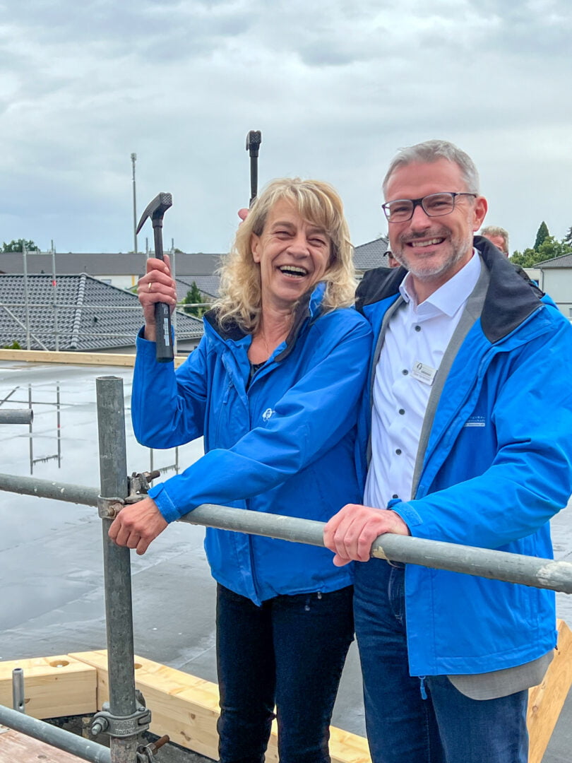 Deckenfest am 4. Juli 2024: Die Vorstände der WBG Otto von Guericke eG stehen auf dem Dach des neuen Anbaus der Geschäftsstelle, halten sich dabei an einem Baugerüst fest und jeweils einen Hammer in der rechten Hand.