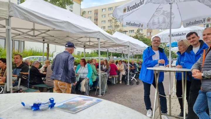 Viele Menschen sitzen unter einem Festzelt und essen und trinken. Rechts im Bild stehen 4 Leute an einem Stehtisch.